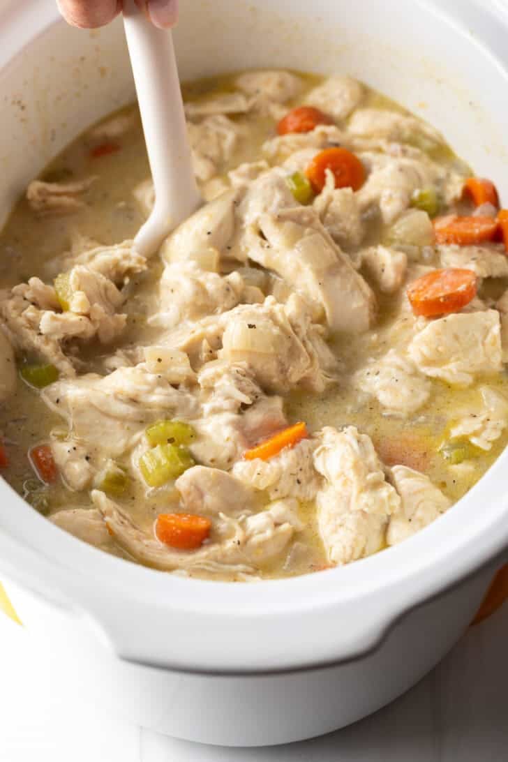 Stirring chicken and dumplings with carrots in a white crock pot.