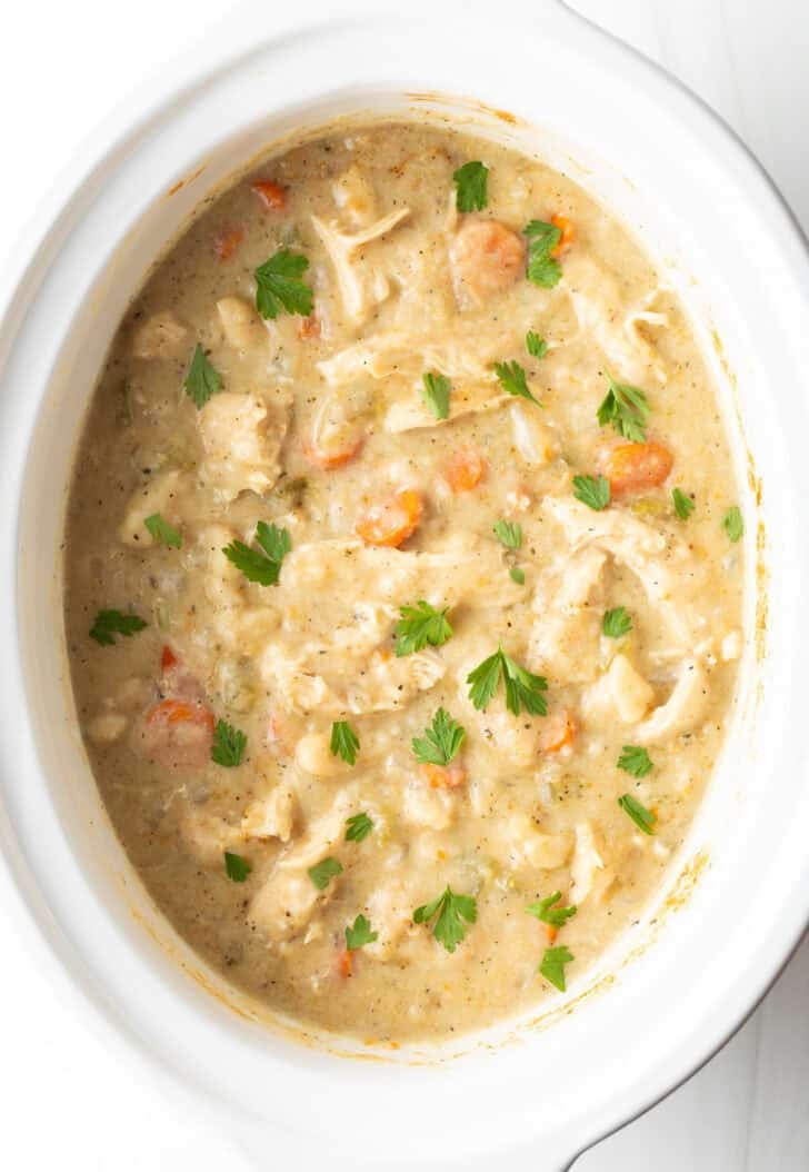 White crock pot with chicken and dumplings sprinkled with fresh parsely.