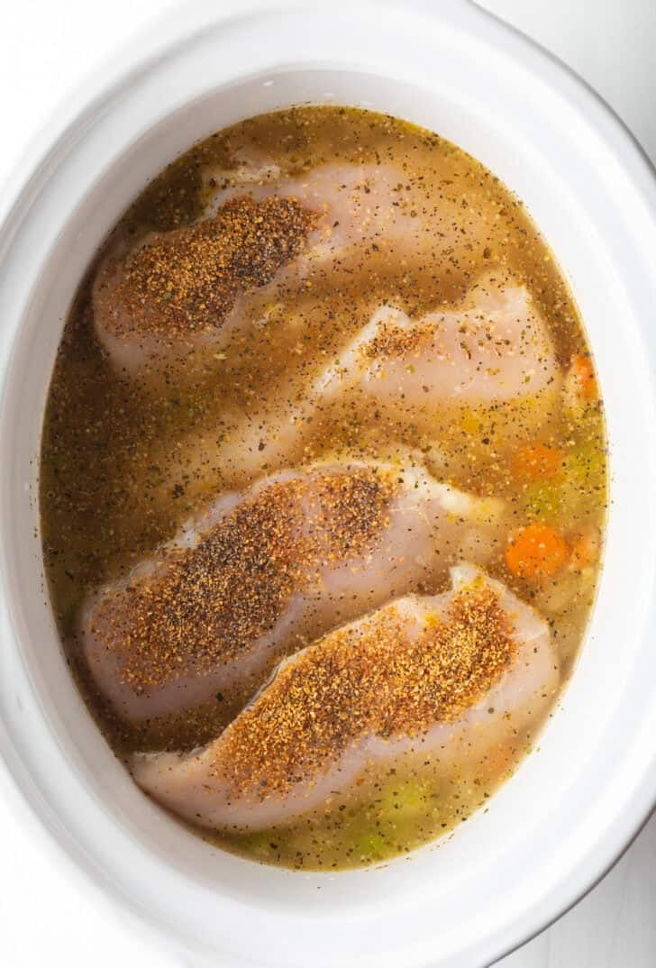 4 raw chicken breasts in a white slow cooker with broth.