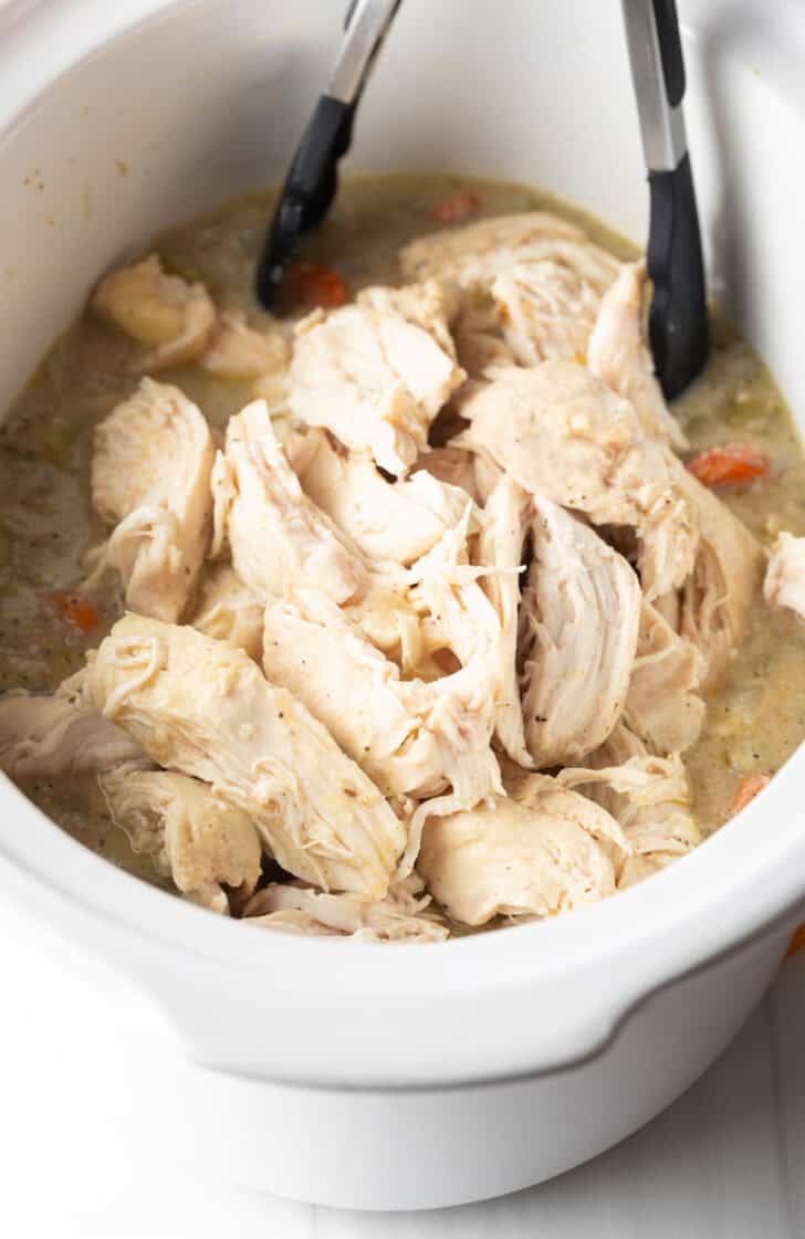 Tongs grabbing shredded chicken from a white crockpot.