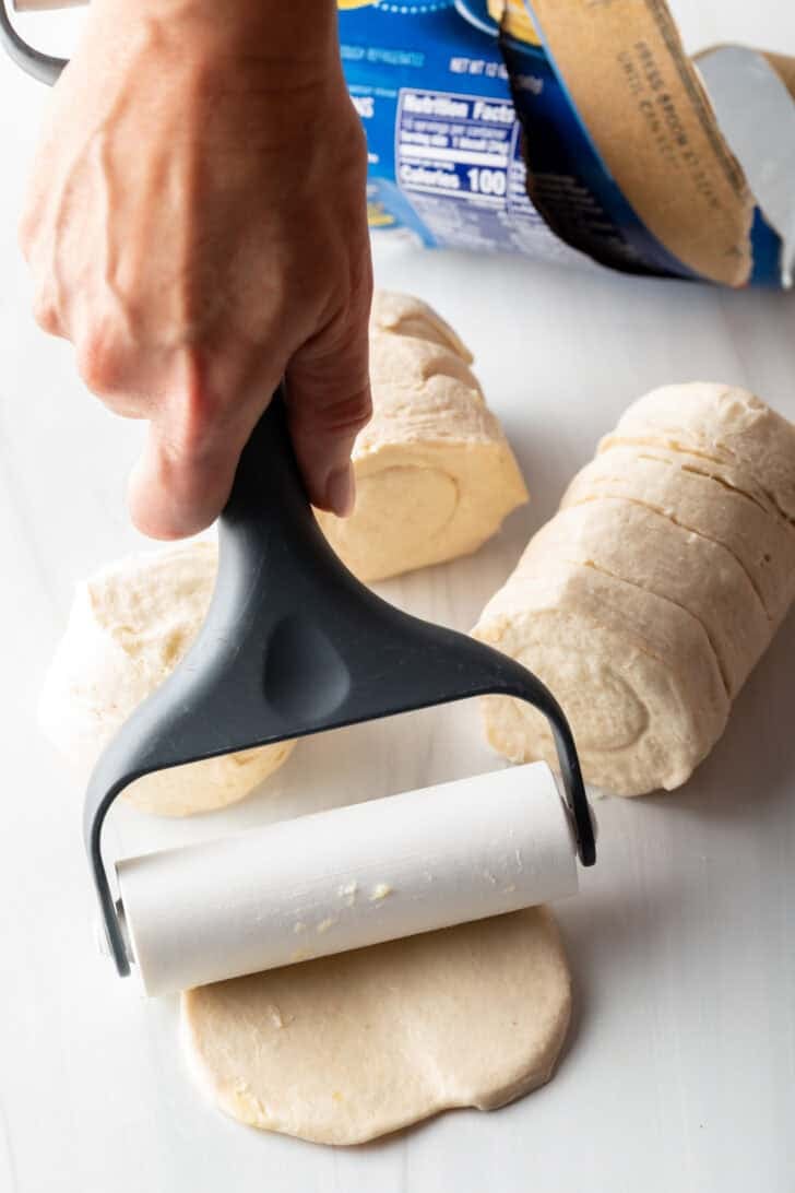 Hand using a dough roller to press a raw biscuit flat.