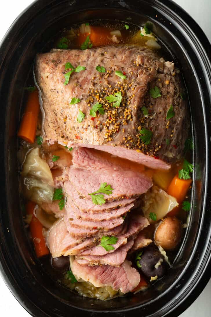 Sliced whole corned beef surrounded by large pieces of carrots, cabbage, and baby potatoes in a black crock pot.