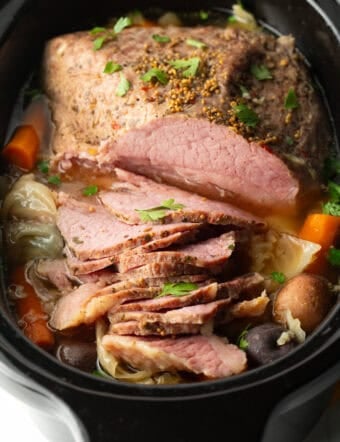 Sliced whole corned beef surrounded by large pieces of carrots, cabbage, and baby potatoes in a black crock pot.