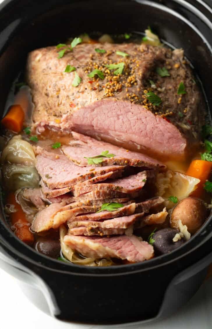 Sliced whole corned beef surrounded by large pieces of carrots, cabbage, and baby potatoes in a black crock pot.