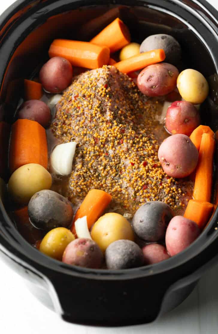 Whole seasoned beef brisket surrounded by raw chunks of carrots and baby potatoes in a black slow cooker.
