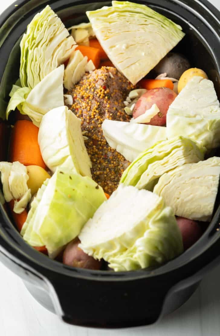 Adding large chunks of cabbage to the seasoned beef brisket surrounded by raw chunks of carrots and baby potatoes in a black slow cooker.