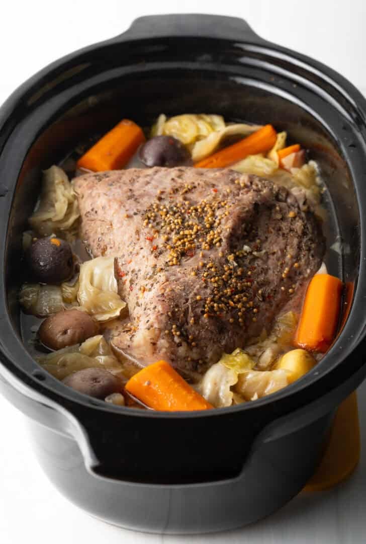 Whole corned beef surrounded by large pieces of carrots, cabbage, and baby potatoes in a black crock pot.