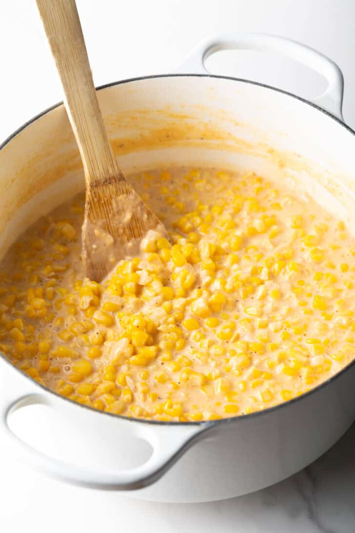 Wood spatula stirring creamed corn in a large white pot.