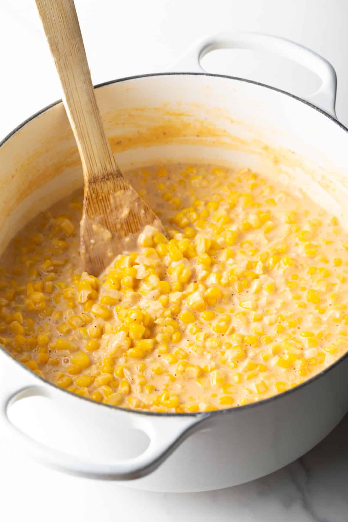 Wood spatula stirring creamed corn in a large white pot.