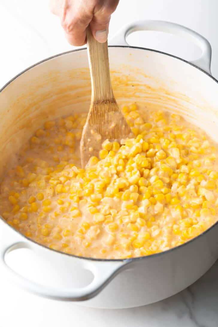 Wood spatula stirring creamed corn in a large white pot.