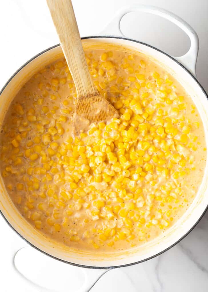 Top down view of a wood spatula stirring our creamed corn recipe in a large white pot.