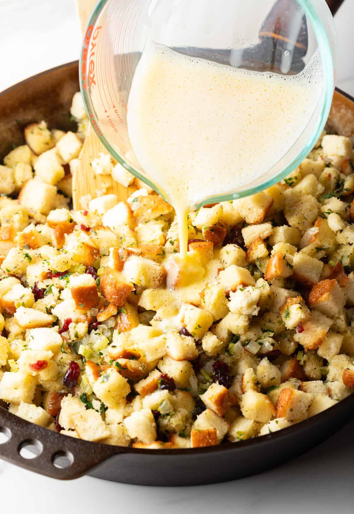 Pouring broth from a glass measuring pitcher overtop of bread cubes in a deep skillet.