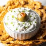White bowl of dirty martini dip topped with one green olive, surrounded by pretzel chips.