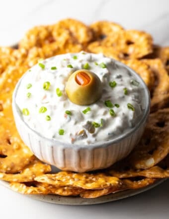 White bowl of dirty martini dip topped with one green olive, surrounded by pretzel chips.