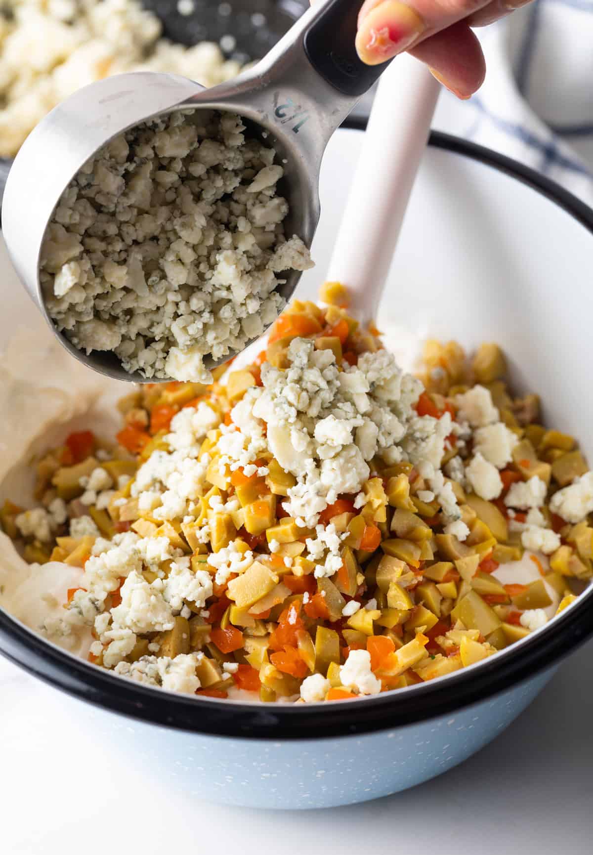 Adding crumbled blue cheese to a bowl of chopped olives, and sour cream and cream cheese.