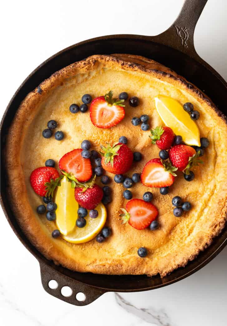 Top down view Dutch baby in a cast iron skillet, topped with strawberries, blueberries, and lemon slices.