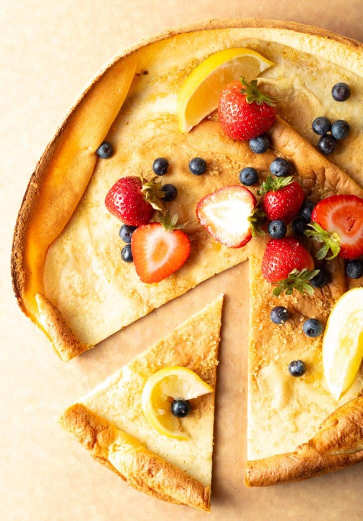 Top down view Dutch baby on a cutting board, topped with strawberries and blueberries, with a slice pulled aside.