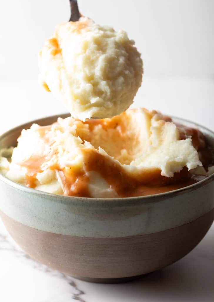 A scoop of mashed potatoes and gravy held over a bowl of mashed potatoes.
