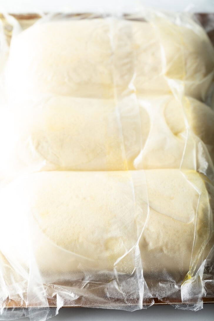 Three loaves of frozen bread dough on a baking sheet covered with plastic.