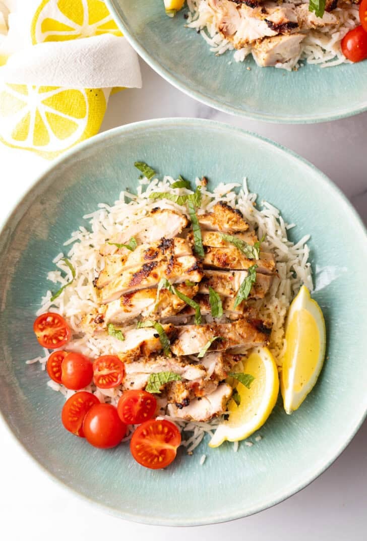 Shallow blue bowl with white rice topped with slices of marinated greek chicken, halved grape tomatoes, and two lemon wedges.