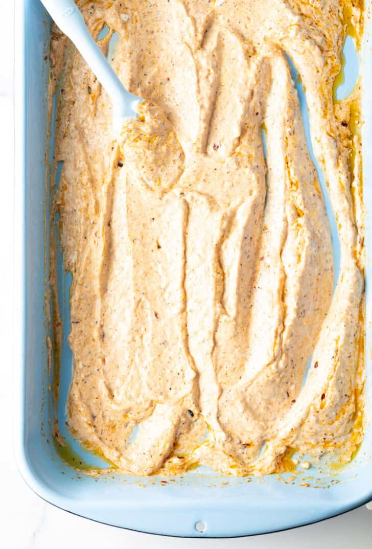 Top down view of a blue spatula spreading yogurt marinade in the bottom of a blue rectangle baking dish.