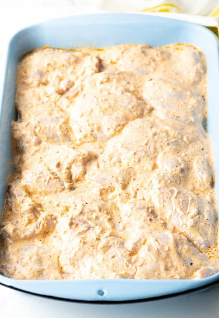 Chicken pieces marinating in a greek yogurt marinade, in a blue rectangle baking dish.
