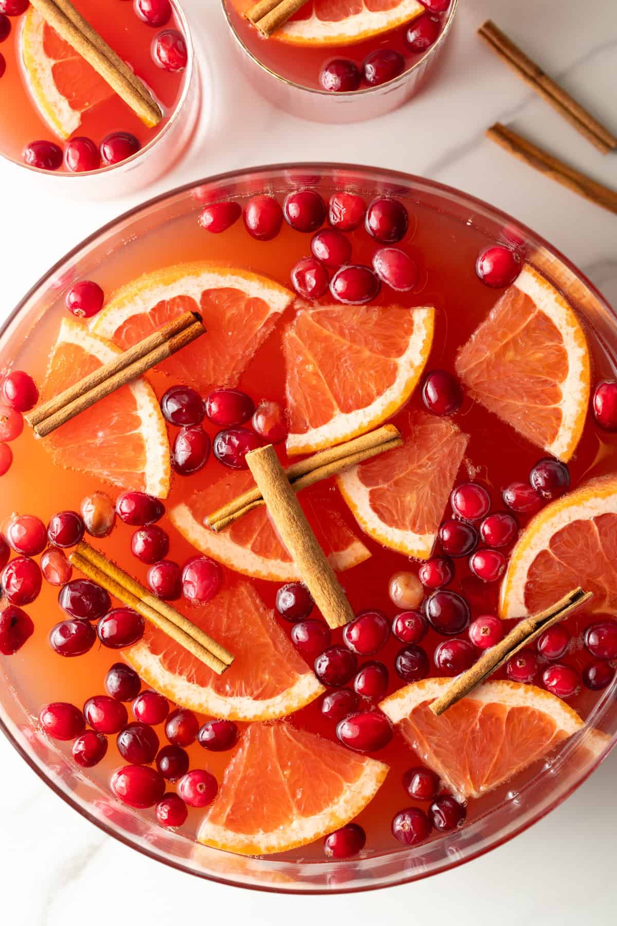 Top down view of a large glass bowlful of dark pink champagne punch with slices of orange, whole cranberries, and cinnamon sticks.