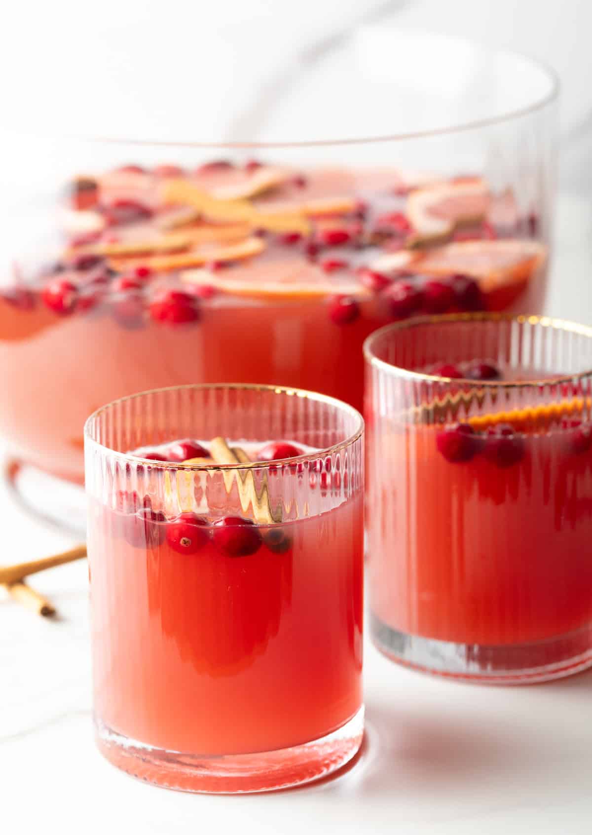 Two glass tumblers of dark pinch holiday punch, garnished with grapefruit slices, whole cranberries, and a cinnamon stick.