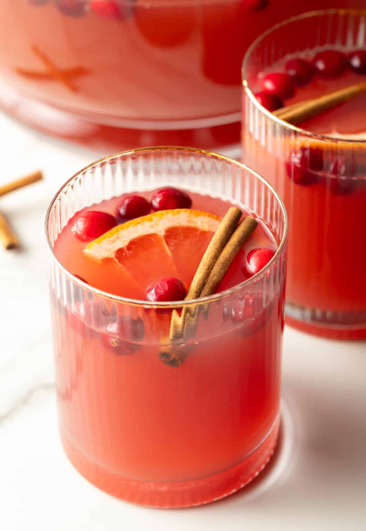 Two glass tumblers of dark pinch holiday punch, garnished with grapefruit slices, whole cranberries, and a cinnamon stick.