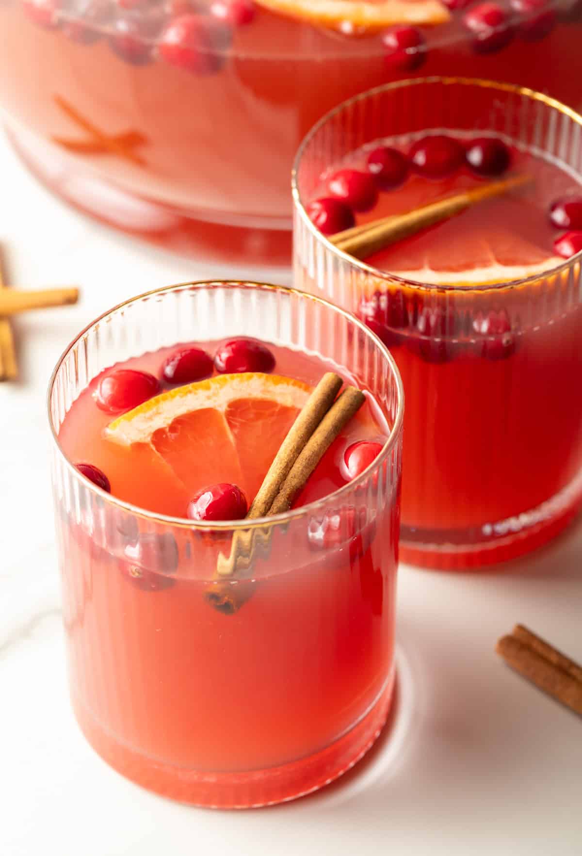 Two glass tumblers of dark pinch holiday punch, garnished with grapefruit slices, whole cranberries, and a cinnamon stick.