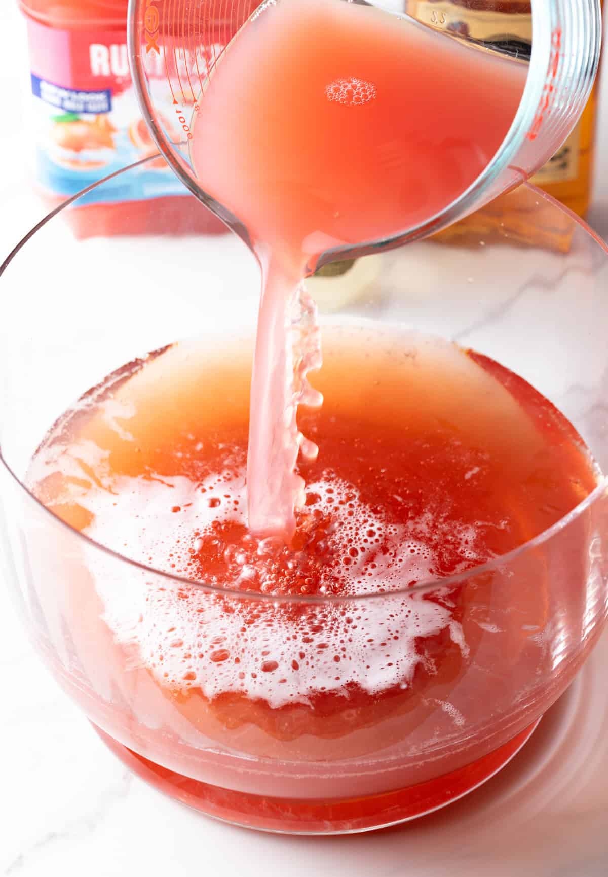 Pouring red grapefruit juice from a glass pitcher into a large glass bowl.