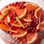 Large glass bowlful of dark pink champagne punch with slices of orange, whole cranberries, and cinnamon sticks.