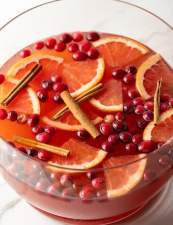 Large glass bowlful of dark pink champagne punch with slices of orange, whole cranberries, and cinnamon sticks.