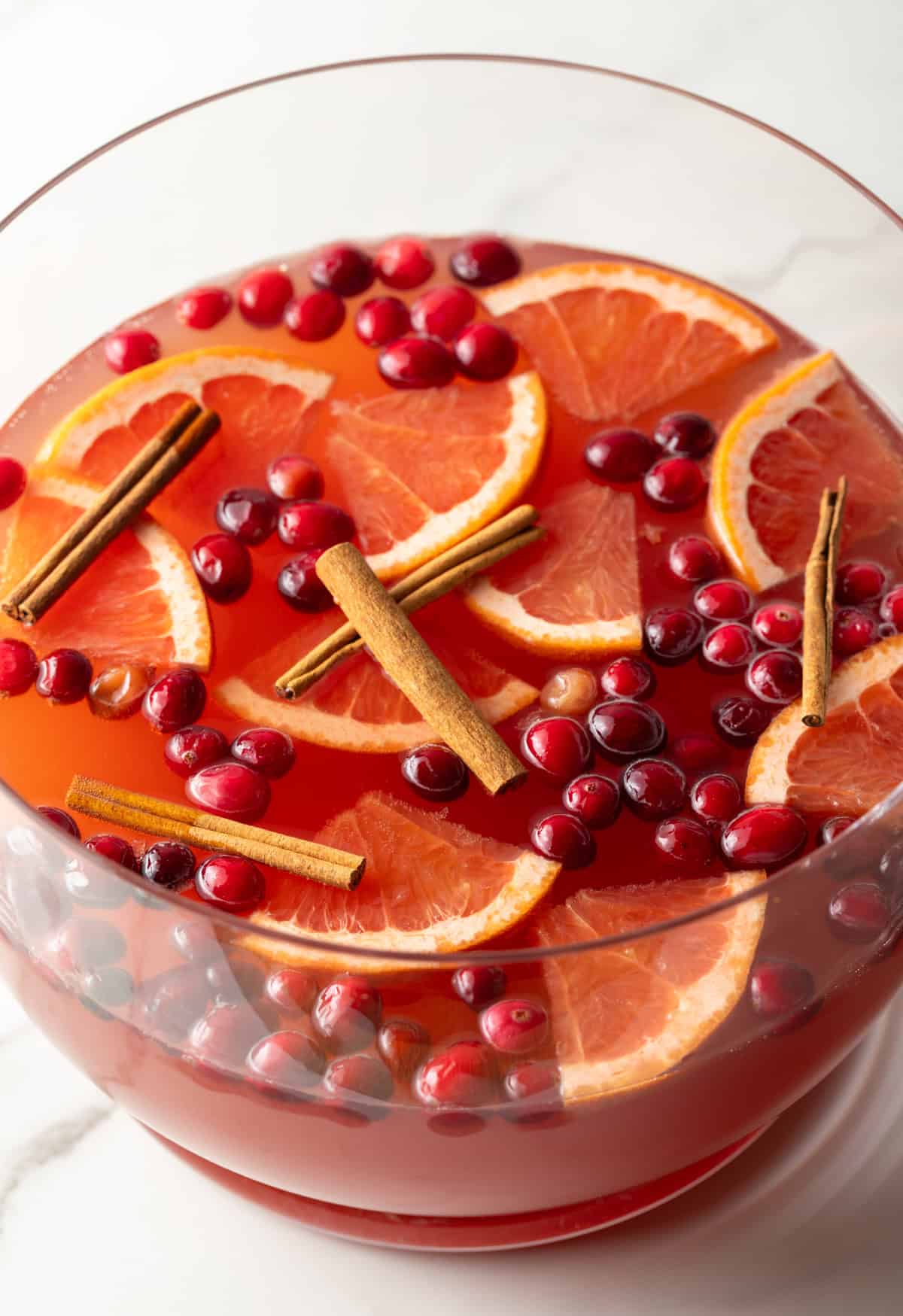 Large glass bowlful of dark pink champagne punch with slices of orange, whole cranberries, and cinnamon sticks.
