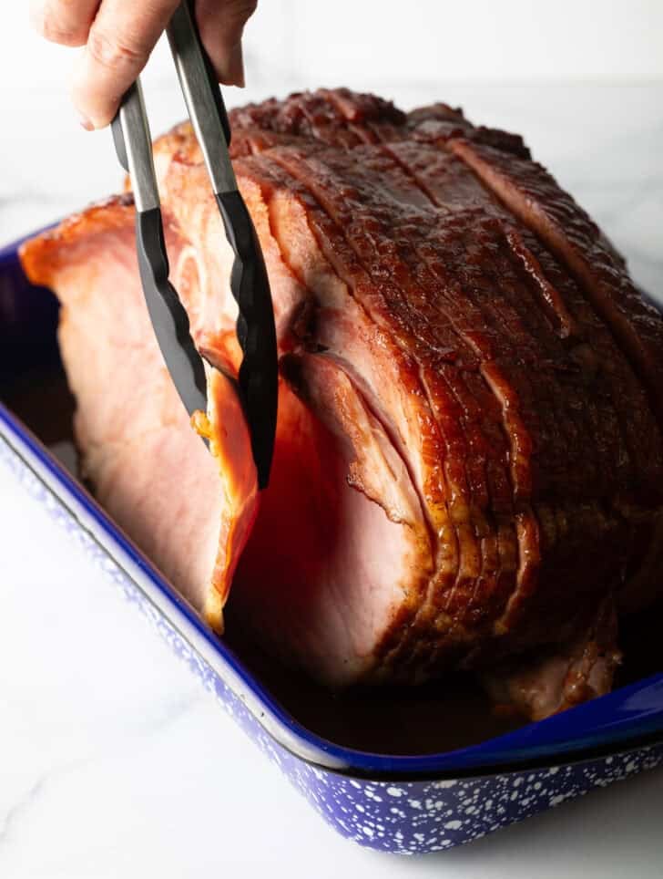 Tongs removing the front slice of a cooked spiral ham in a baking dish.