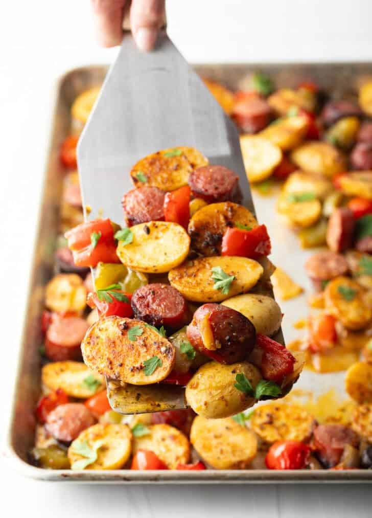 Metal spatula holding up a scoop of kielbasa and potatoes with red peppers.