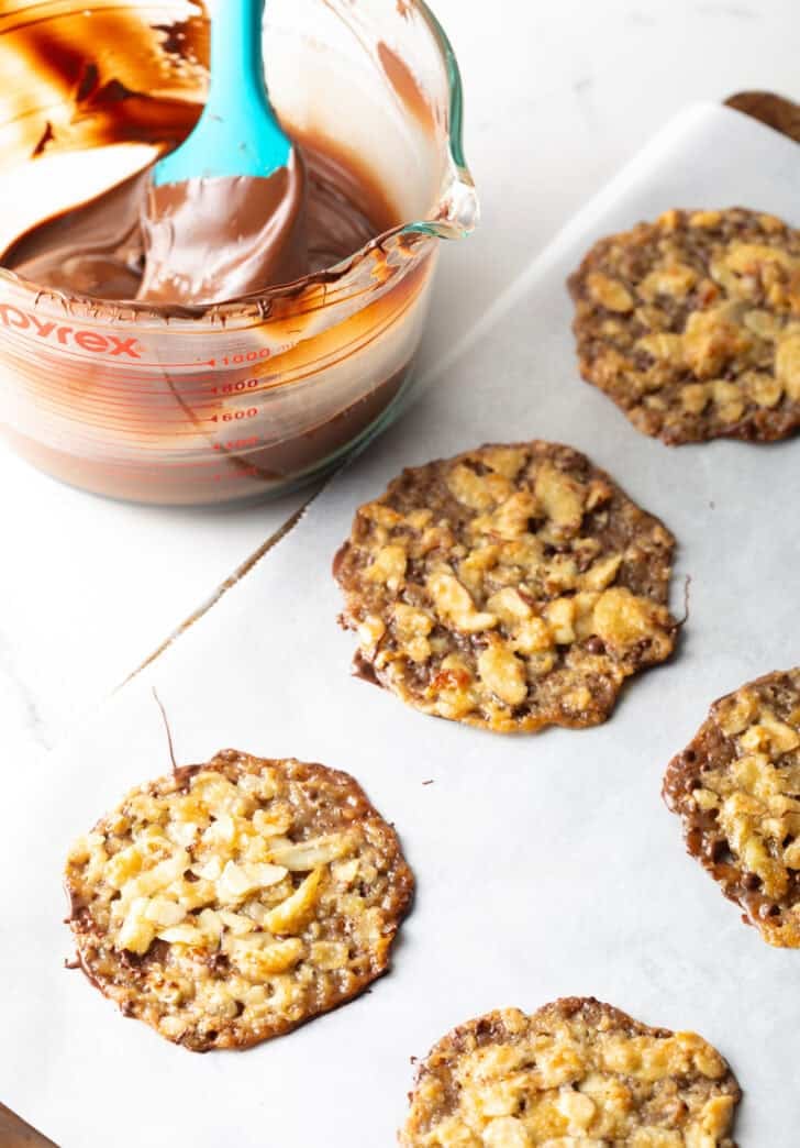 Lace cookies on parchment paper next to a glass bowl full of melted chocolate.