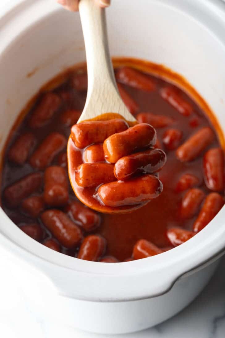 Wooden spoonful of smoked mini hotdogs in bbq sauce, held over the crock pot to camera.