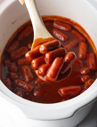 Wooden spoonful of smoked mini hotdogs in bbq sauce, held over the crock pot to camera.