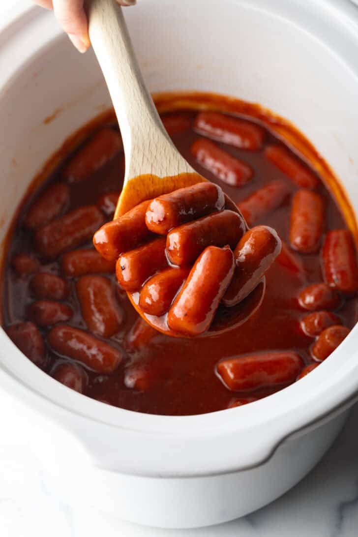 Wooden spoonful of smoked mini hotdogs in bbq sauce, held over the crock pot to camera.