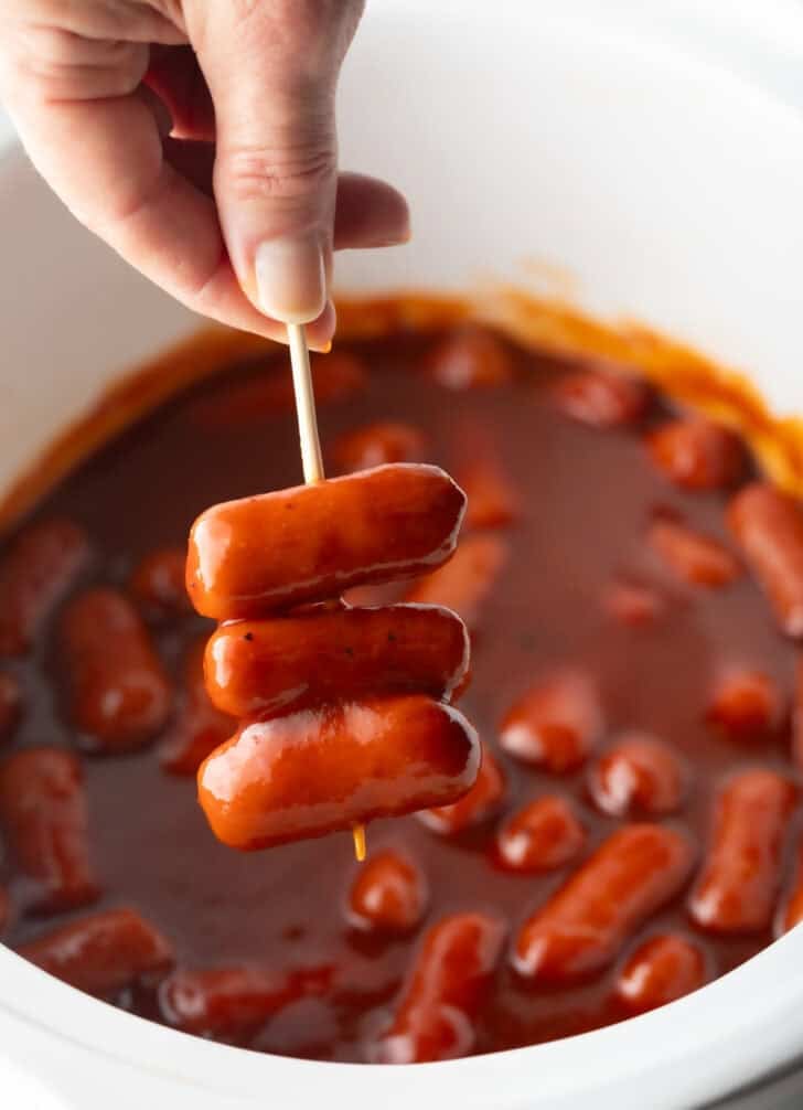 Hand holding a skewer with 3 little smokies, over a crock pot with cooked mini sausages in bbq sauce.