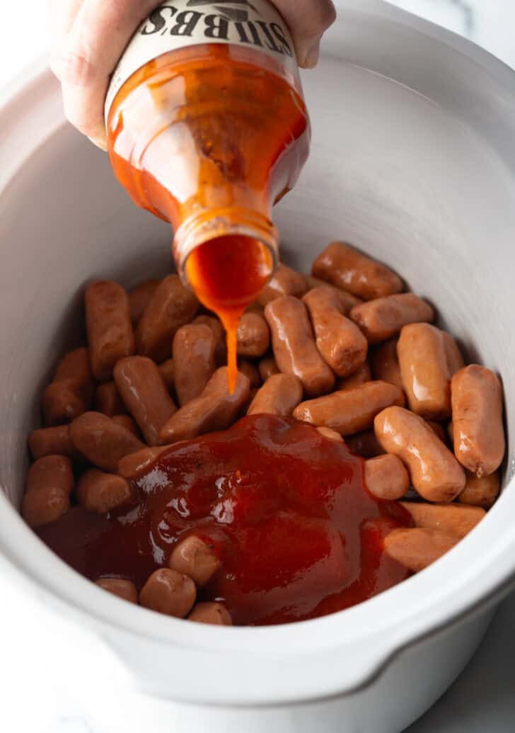 Pouring BBQ sauce over lil smokies in a white crock pot.