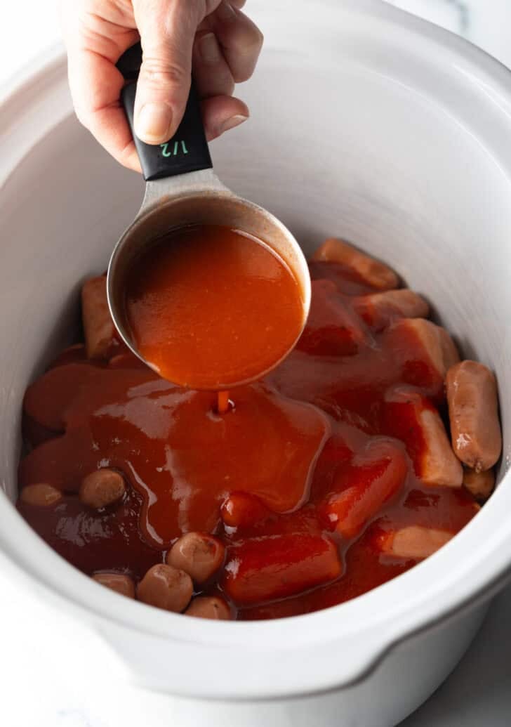 Hand pouring hot sauce from a measuring cup into a slow cooker with smokies.