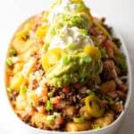 Large white oval serving dish loaded with tater tot nachos with ground beef, jalapenos, salsa, cheddar cheese, guacamole and sour cream.