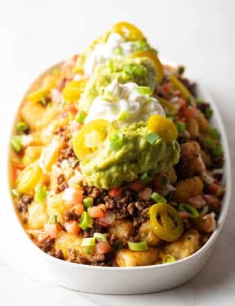 Large white oval serving dish loaded with tater tot nachos with ground beef, jalapenos, salsa, cheddar cheese, guacamole and sour cream.