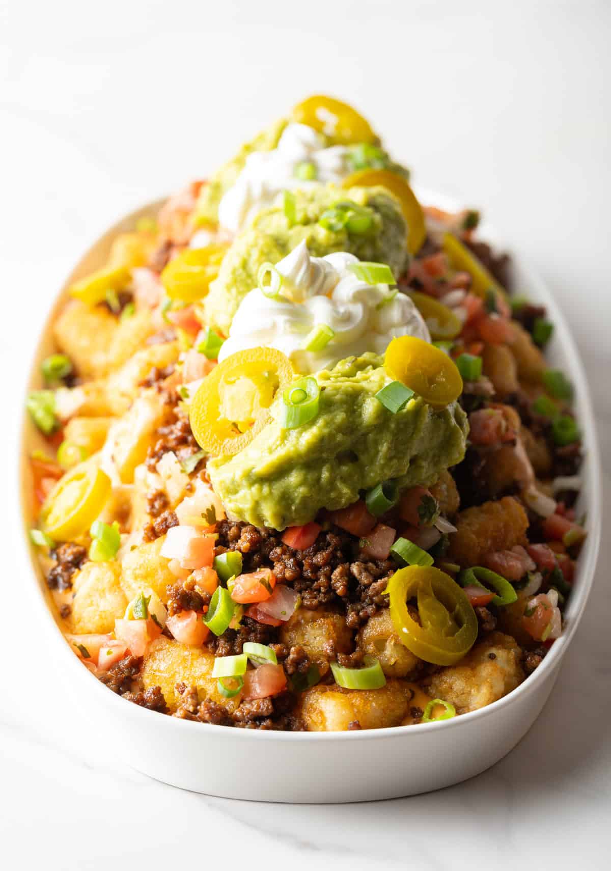 Large white oval serving dish loaded with tater tot nachos with ground beef, jalapenos, salsa, cheddar cheese, guacamole and sour cream.