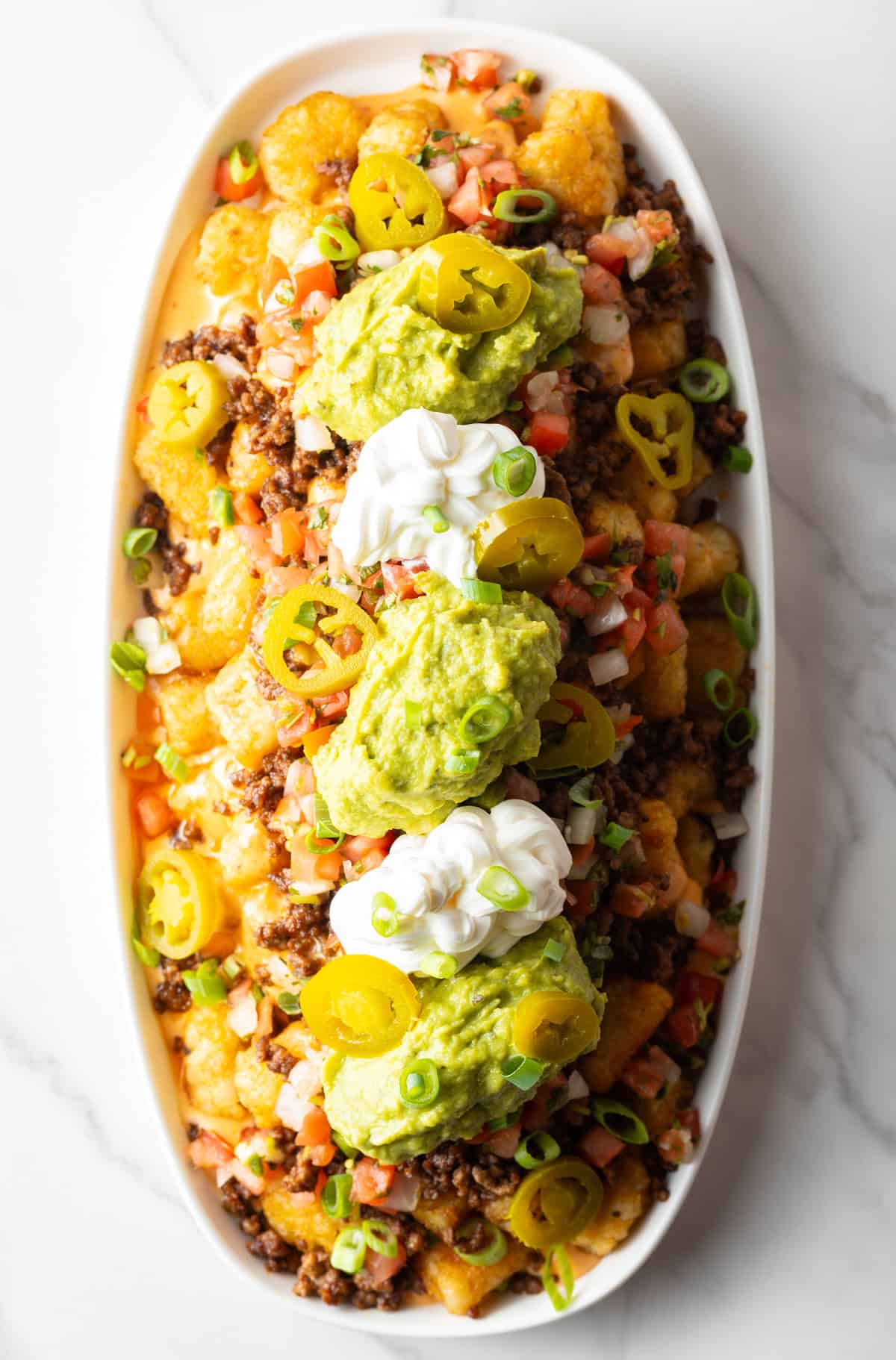 Top down view of large white oval serving dish loaded with tater tot nachos with ground beef, jalapenos, salsa, cheddar cheese, guacamole and sour cream.