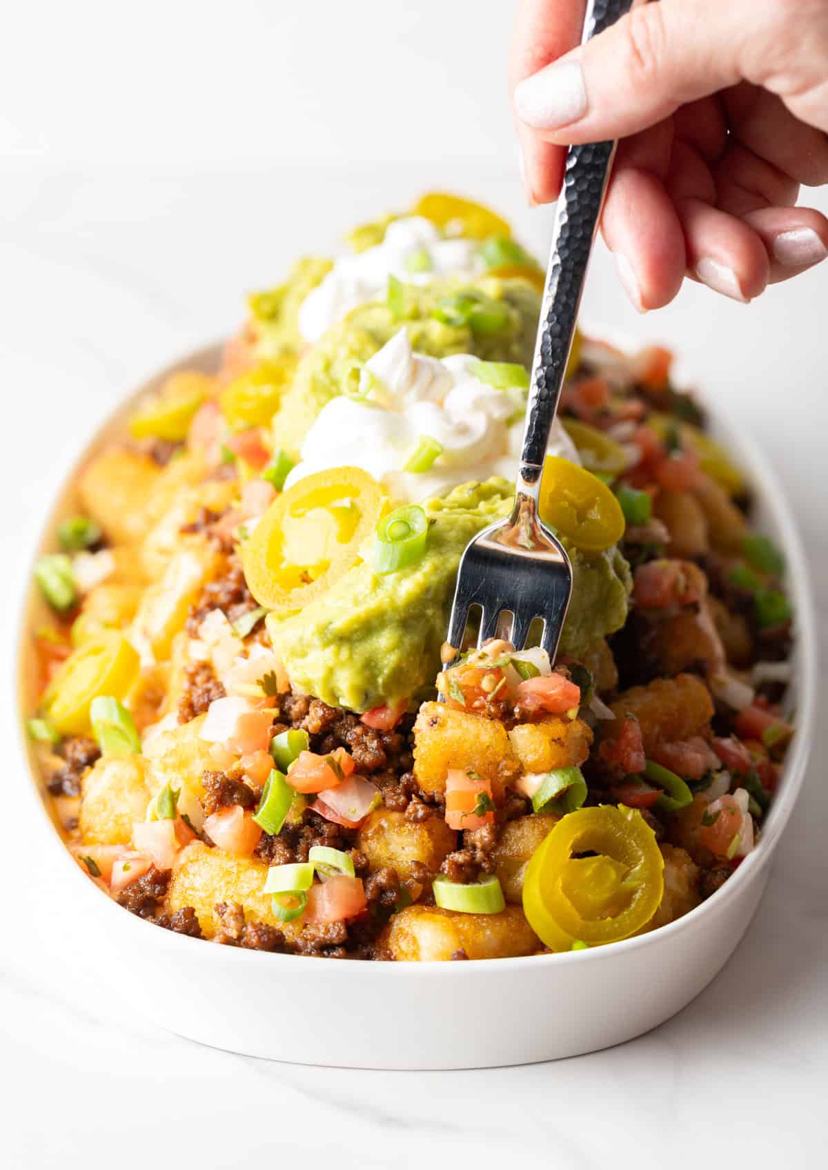 Hand with a fork spearing a bite of totchos from a large white oval serving dish.