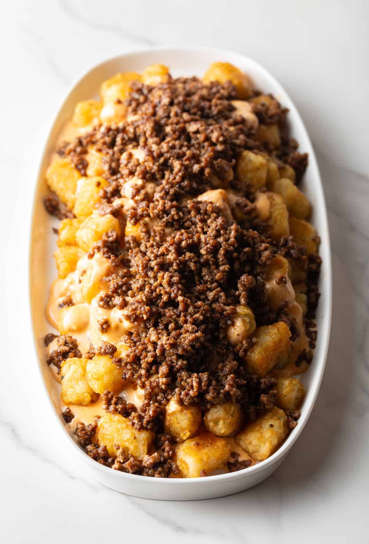 Tater tots in a large white oval bowl covered with ground beef.