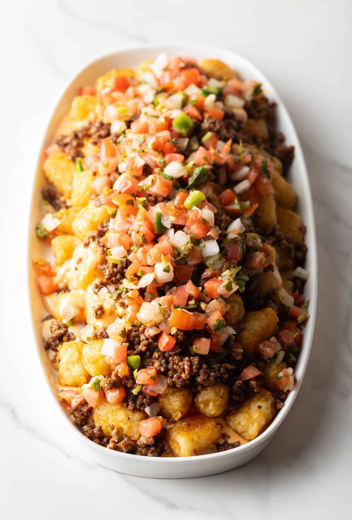 Tater tots in a large white oval bowl covered with ground beef and pico de gallo.